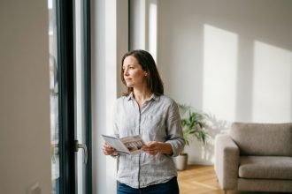 Femme examinant sa fenêtre ancienne avant de choisir un remplacement, guide travaux à la main dans un salon moderne
